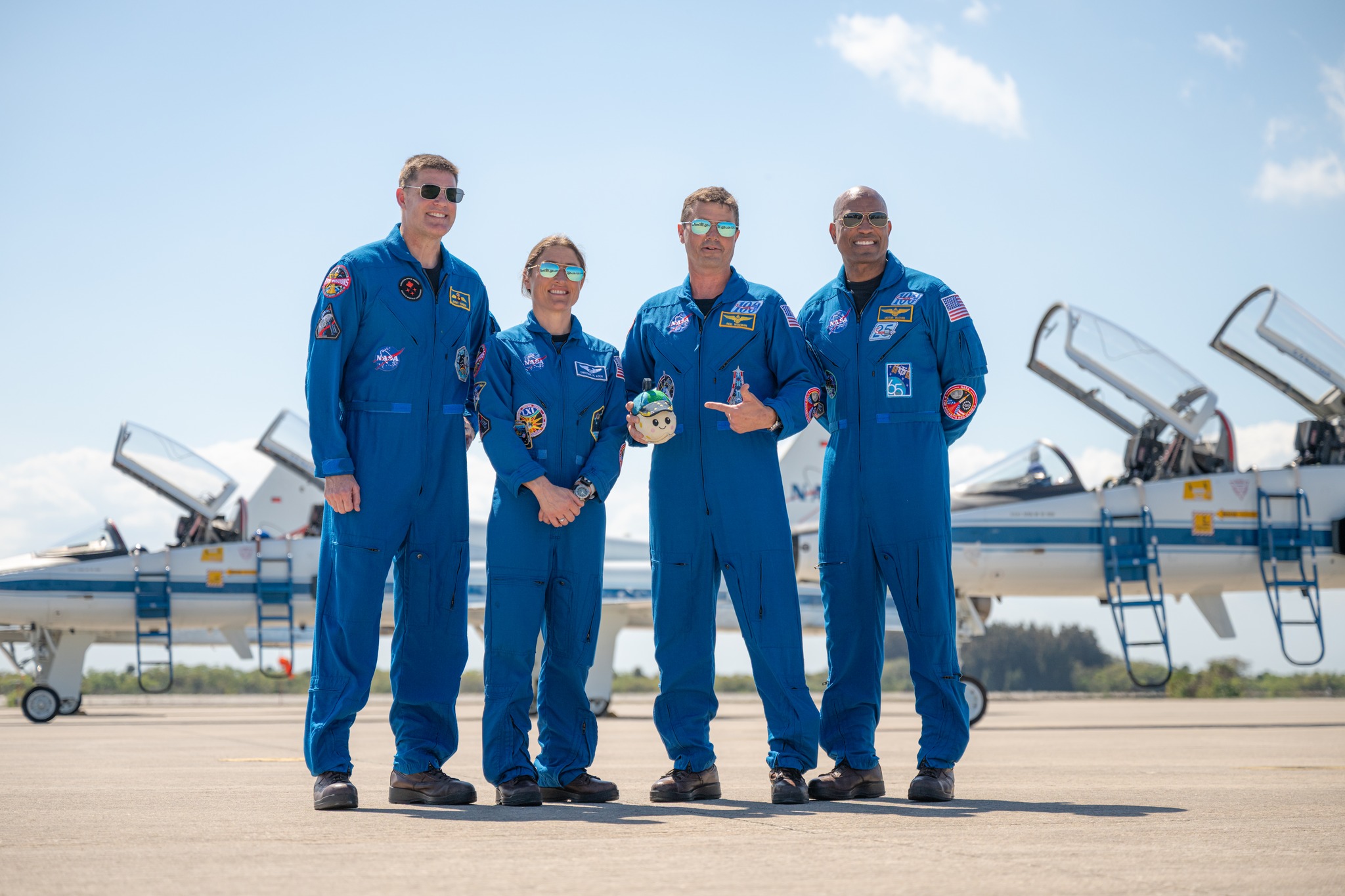 The four Artemis II astronauts standing on the tarmac with fighter jets behind them. They are wearing blue flight uniforms and sunshades.