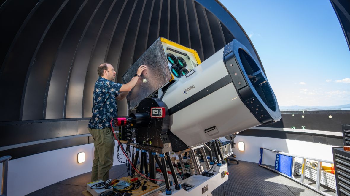 A man stands next to a relatively large optical telescope, and is looking into the side of it. The telescope is located inside the dome of an observatory which is open.