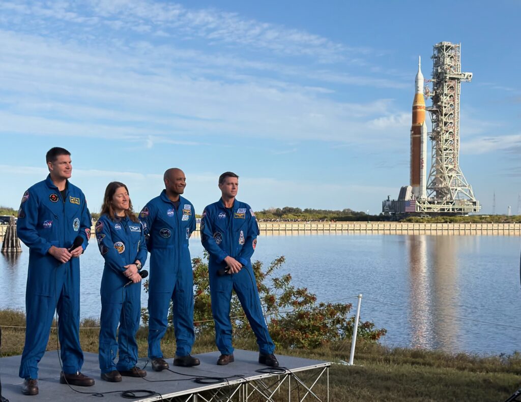 The four Artemis II astronauts stand on a media platform, with the Artemis II rocket in the background.