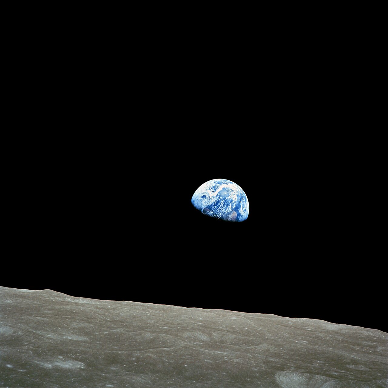 The Earth, suspended in space in the distance, whilst a portion of the lunar surface is in the foreground. The Earth is bright and blue in a sea of darkness and the Moon is grey and desolate.