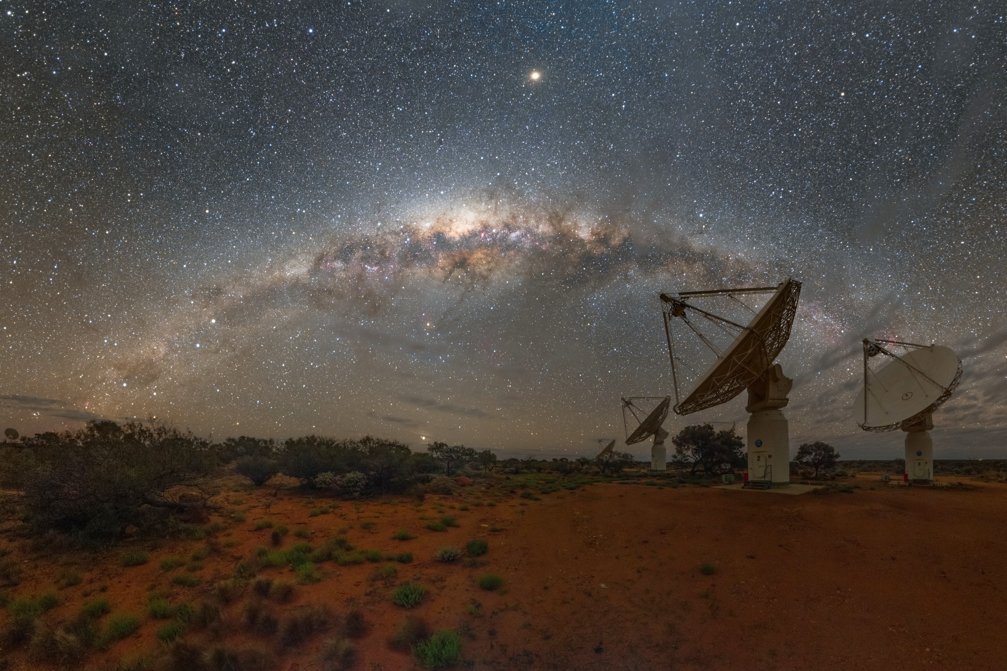 Four radio dish antennas on the right hand side of the image. The ground below them is the red earth sands and gravel of the Australian outback. In the sky, the Milky Way arches overhead.